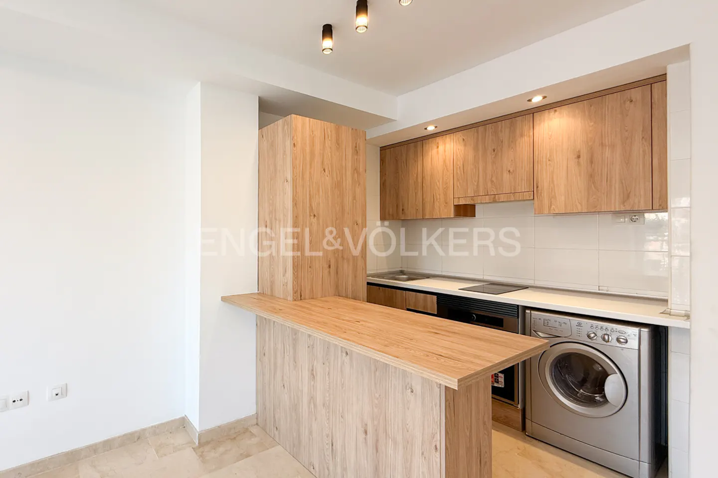 Bright kitchen with wood cabinets, white walls, and stainless steel washing machine. A breakfast bar extends from the cabinets.