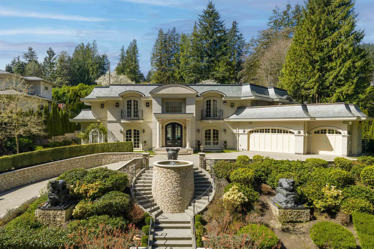 Beige two-story house with a gray roof, a stone fountain, and green landscaping.