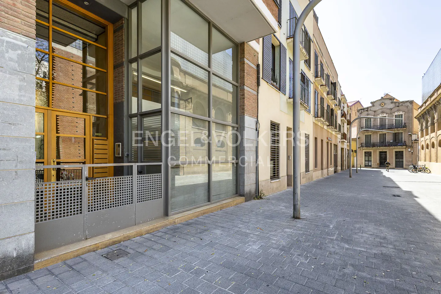 Exterior view of a commercial building with large glass windows and a gray metal gate on a cobblestone street.