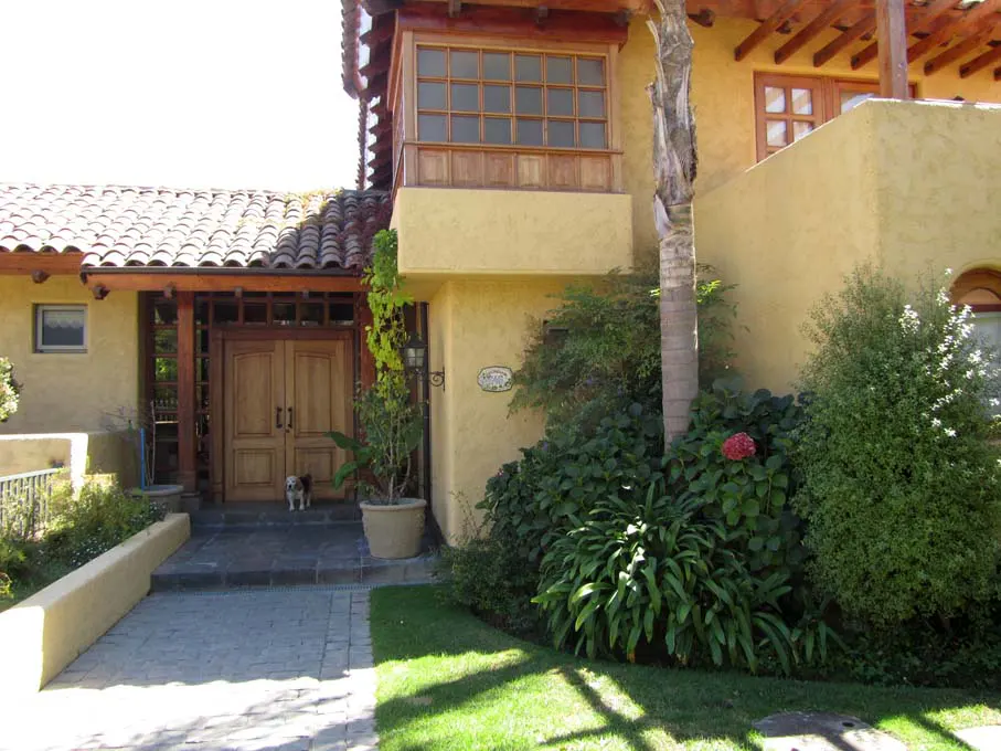 Exterior of a yellow stucco house with a red tile roof, wooden doors, and lush green landscaping. A small dog stands near the entrance.