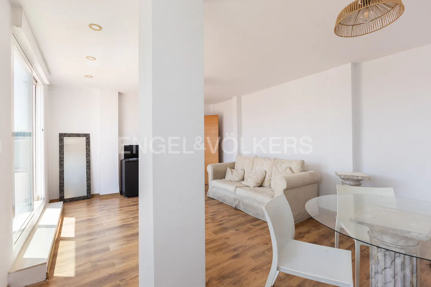 Bright living room with wood floors, white walls, and a beige sofa. A glass table with column-style legs sits near white chairs. A mirror and cabinet are visible.