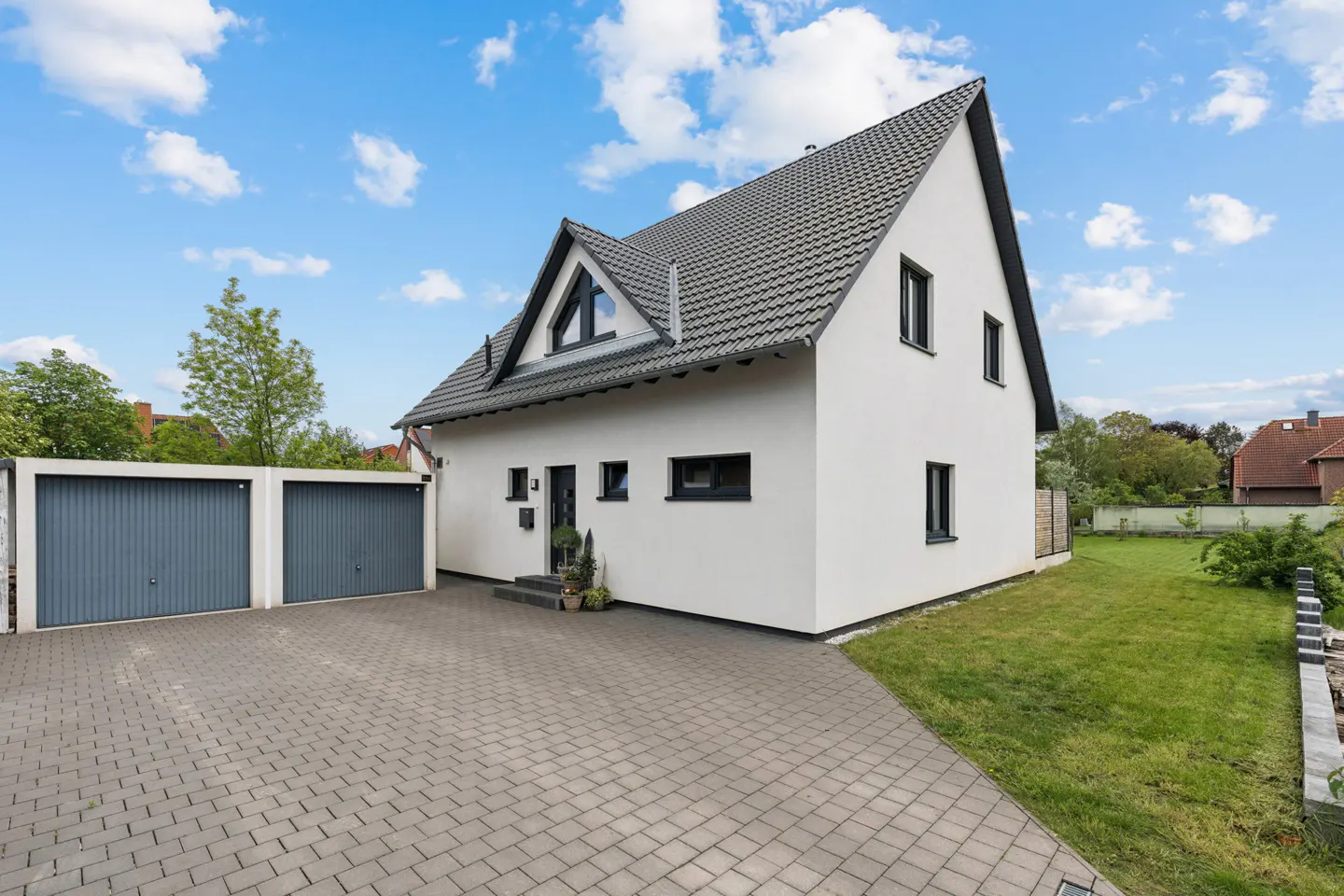 Modern white house with a dark grey roof, double garage, and paved driveway under a blue sky with scattered clouds.