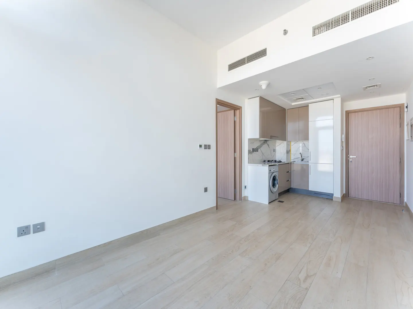 Bright, empty apartment with light wood floors, white walls, and a compact kitchen with gray and white cabinets. A washing machine is visible.