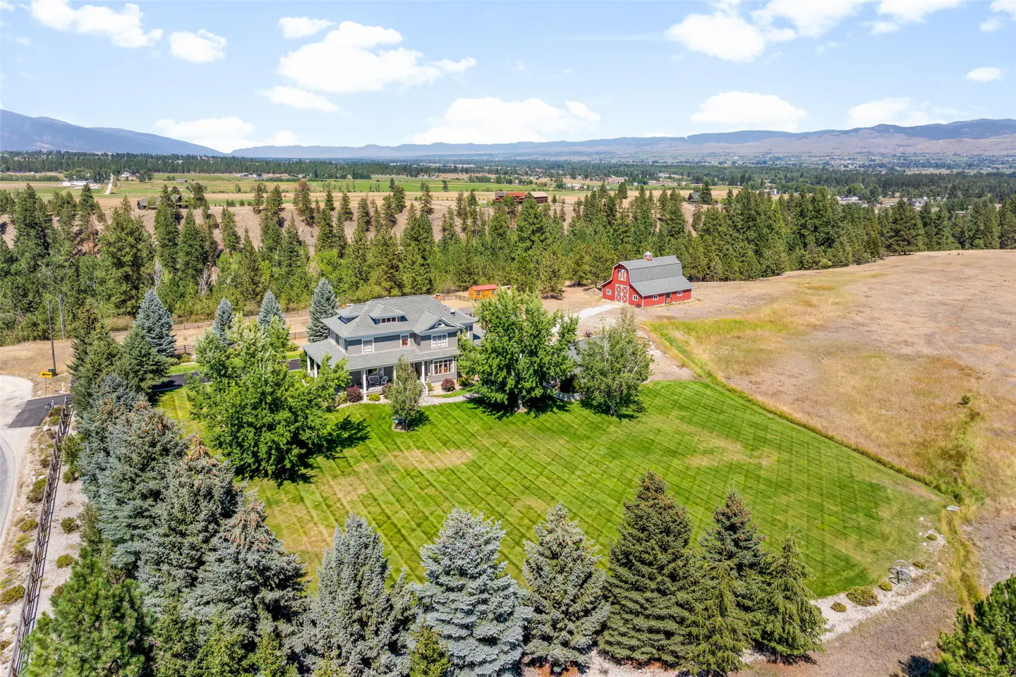 Aerial view of a gray house with a red barn, surrounded by green trees and a large lawn. Mountains are visible in the background.
