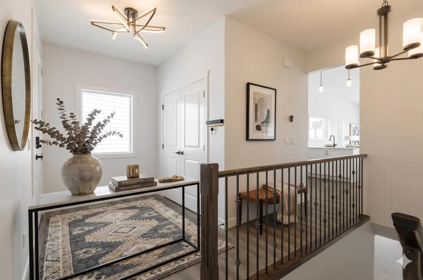 Bright hallway with white walls, a table with a vase of dried flowers, and a patterned rug. A staircase with a dark wood railing leads to the lower level.