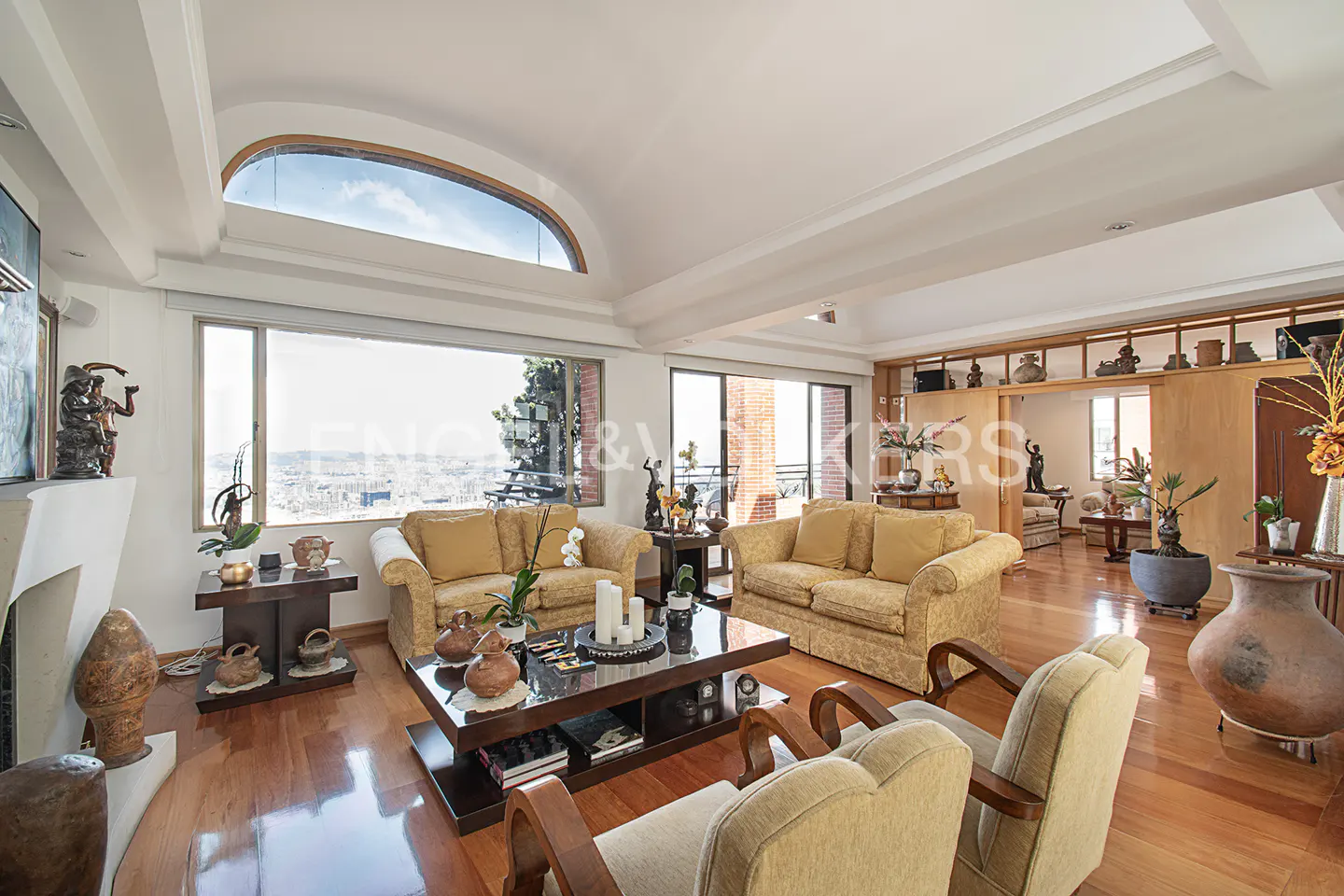 Bright living room with hardwood floors, tan sofas, and a large window overlooking a city view.