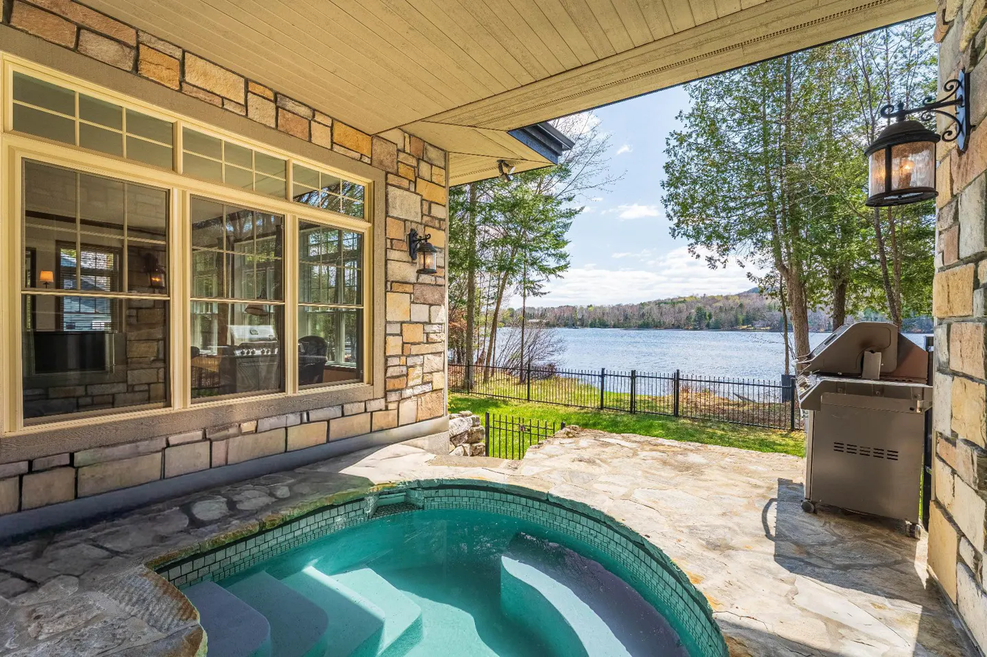 Outdoor patio with a turquoise hot tub, stone flooring, and a stainless steel grill overlooking a lake.