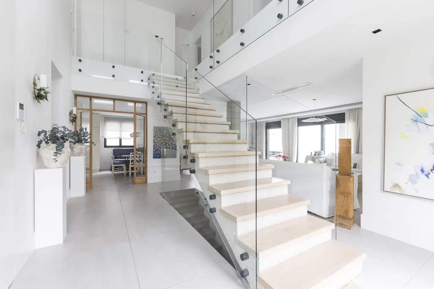 Bright, modern home interior with white walls, light wood stairs, and glass railings. A living room and dining area are visible in the background.