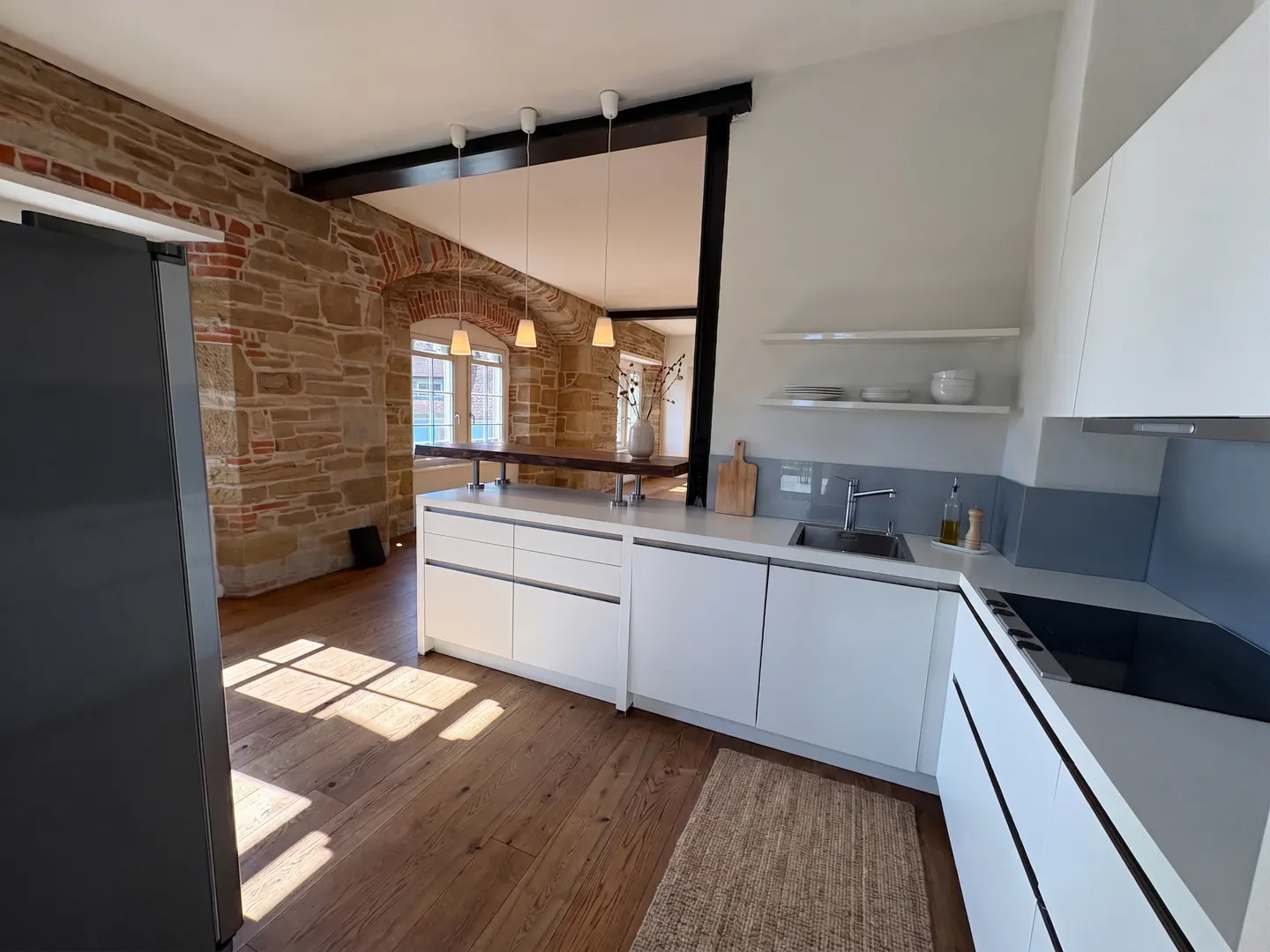 Modern kitchen with white cabinets, wood floors, and exposed brick wall. A dining table is visible through an opening.
