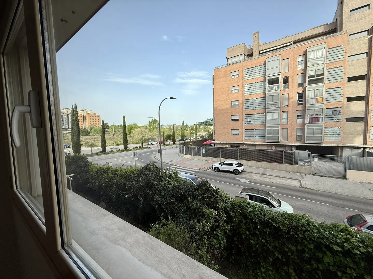 View from a window overlooking a street with cars, trees, and a brick apartment building under a blue sky.