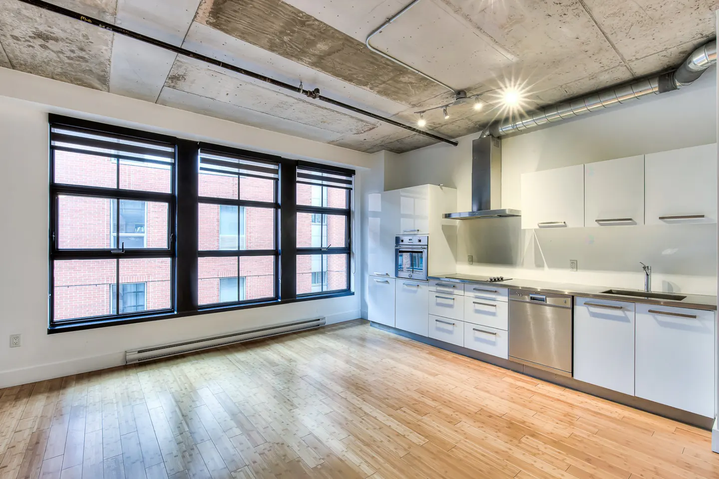 Bright, modern apartment with hardwood floors, white cabinets, and large windows with black frames. Exposed concrete ceiling.