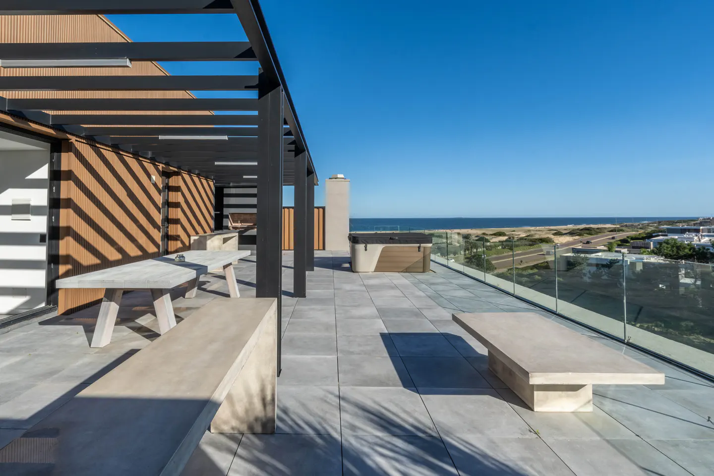 Rooftop patio with ocean view. Gray tile floor, concrete benches and tables, black pergola, and a hot tub. Clear blue sky.