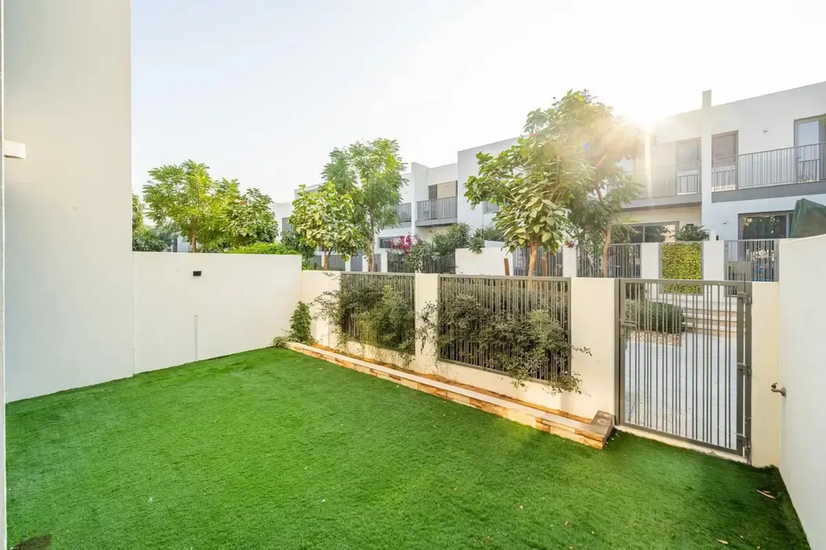 A backyard with green grass, white walls, and a metal gate leading to other houses.