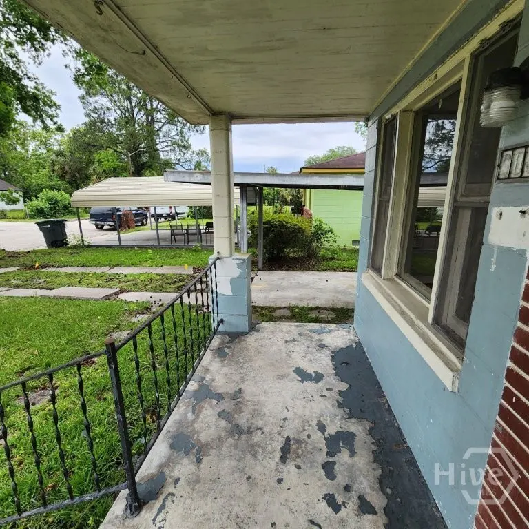 Exterior view of a home's porch with a black railing, peeling blue paint, and a view of the yard and carport.
