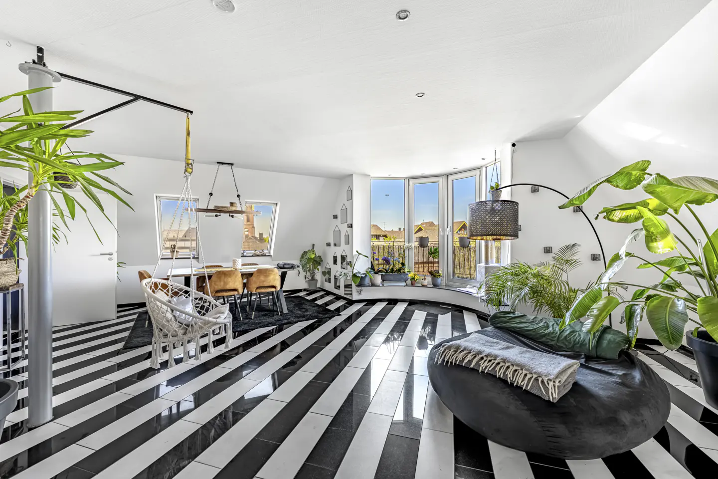 Bright, modern living space with black and white striped floors, plants, and a hanging chair. A dining table sits near a window.