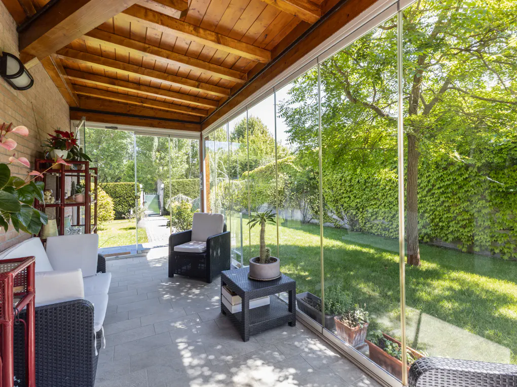 Sunroom with glass walls overlooking a green lawn. Wicker furniture and a wood ceiling create a cozy space.