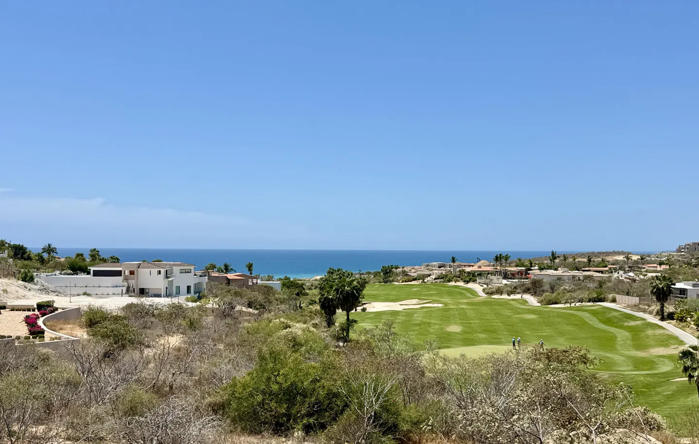Scenic view of a golf course and ocean. Green fairway, blue water, and clear sky. Houses and desert vegetation surround the course.