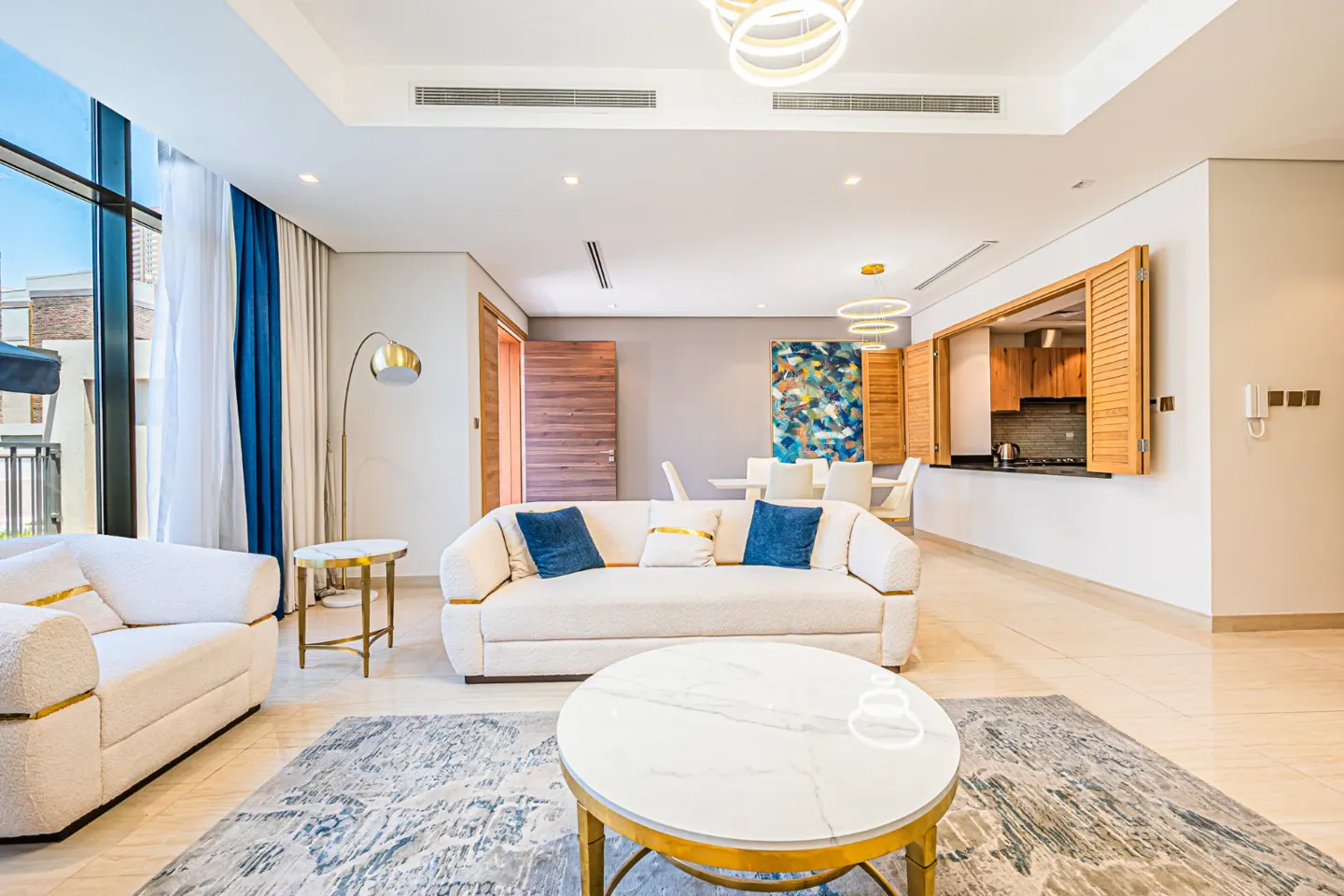 Bright living room with white sofas, blue pillows, and a marble coffee table on a patterned rug. A kitchen pass-through is visible.