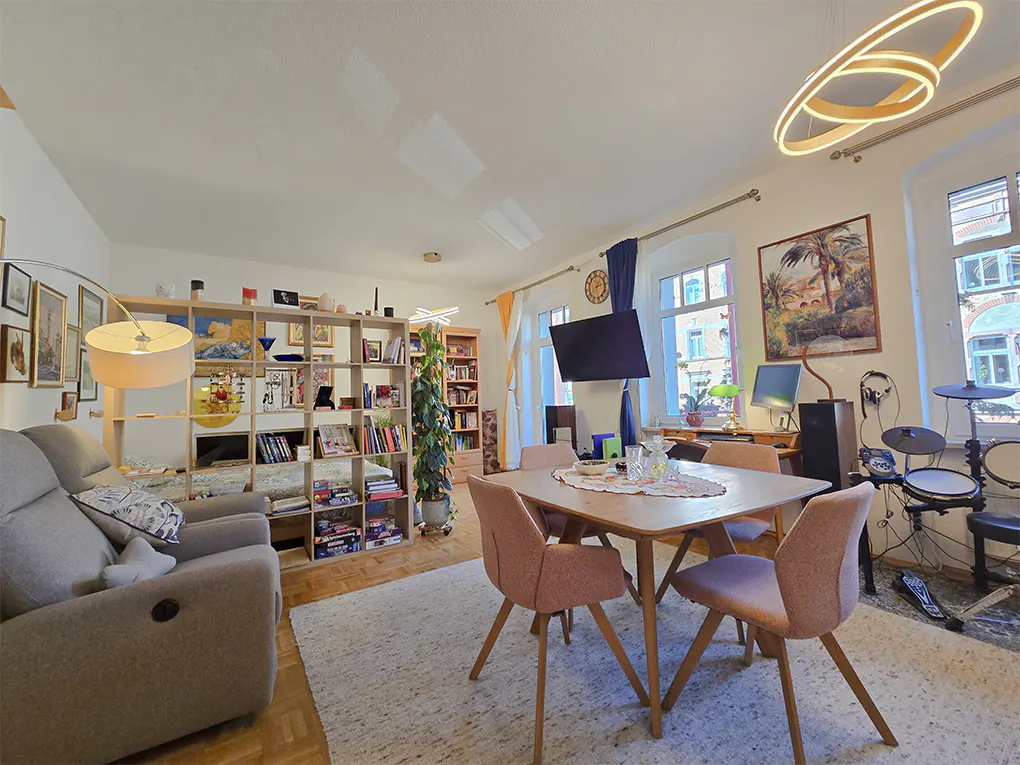 Bright living room with a dining table, pink chairs, a gray sofa, and a bookshelf filled with books and decorations. A drum set is visible in the background.