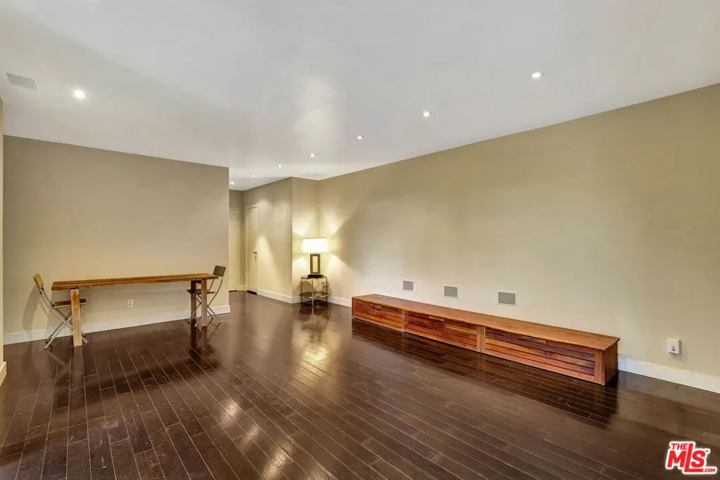 An interior shot of a room with dark wood floors, beige walls, and recessed lighting. A long wooden table with two chairs sits on the left, and a long wooden cabinet is on the right.