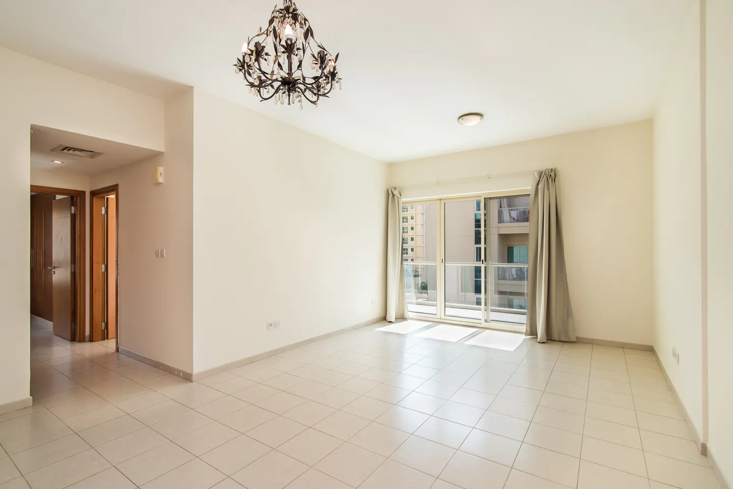 Empty room with white walls, tiled floor, and a chandelier. Sliding glass doors lead to a balcony with a city view.