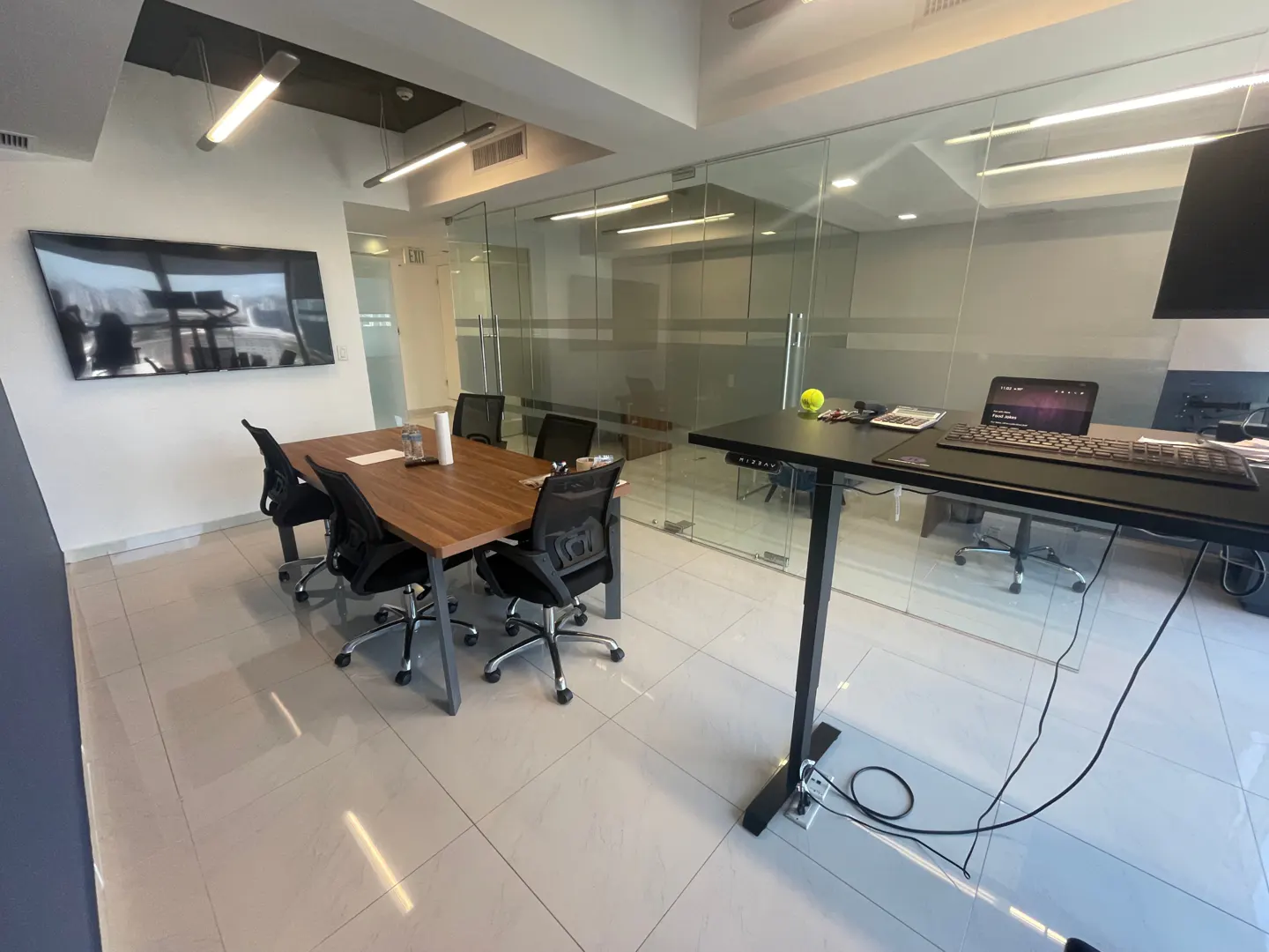 Office space with a wood conference table, black chairs, and a large screen TV on the wall. A standing desk with a computer is nearby.