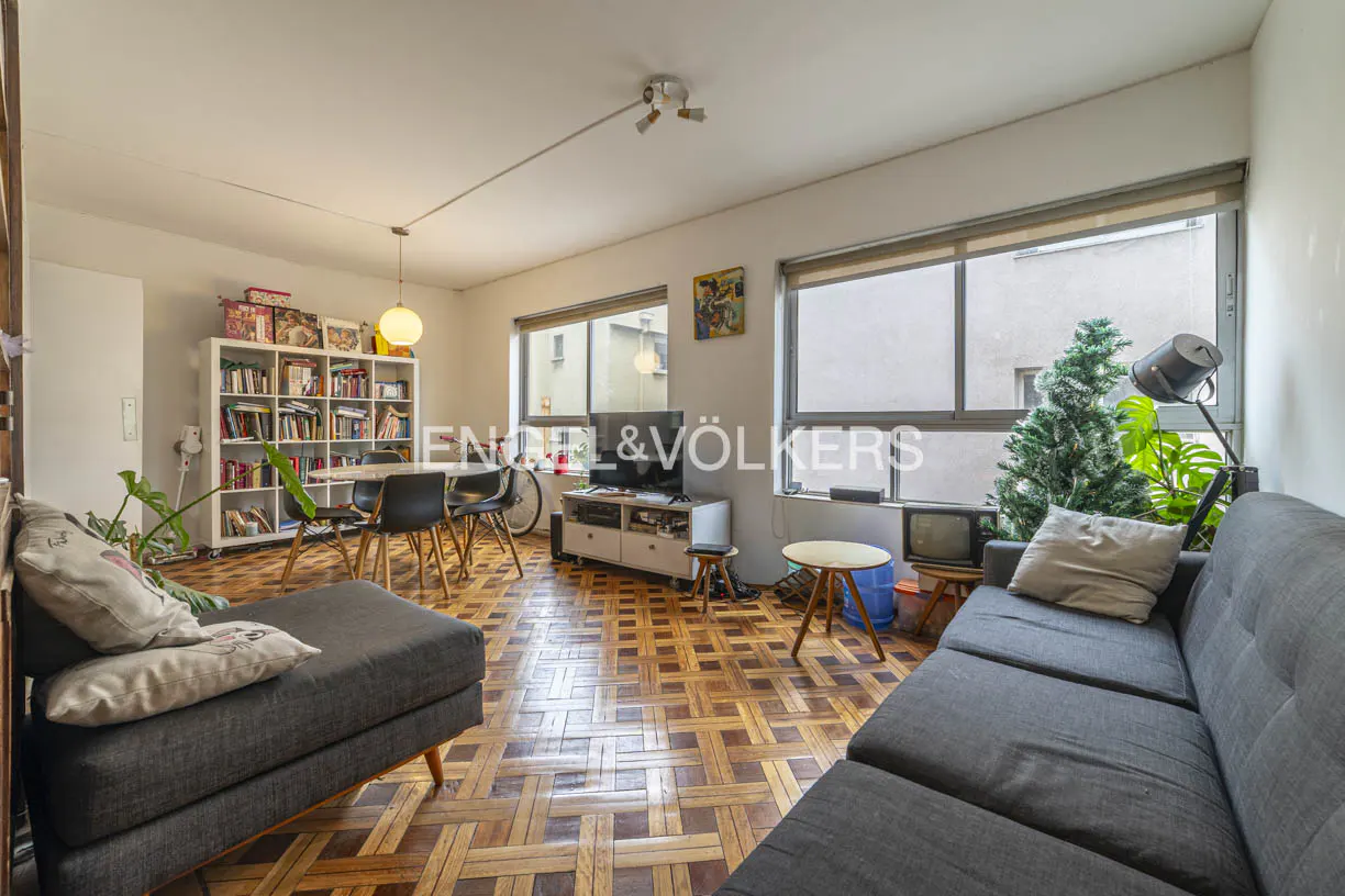 Living room with parquet floors, grey sofa, dining area with bookshelf, TV stand, and large windows with a small tree.