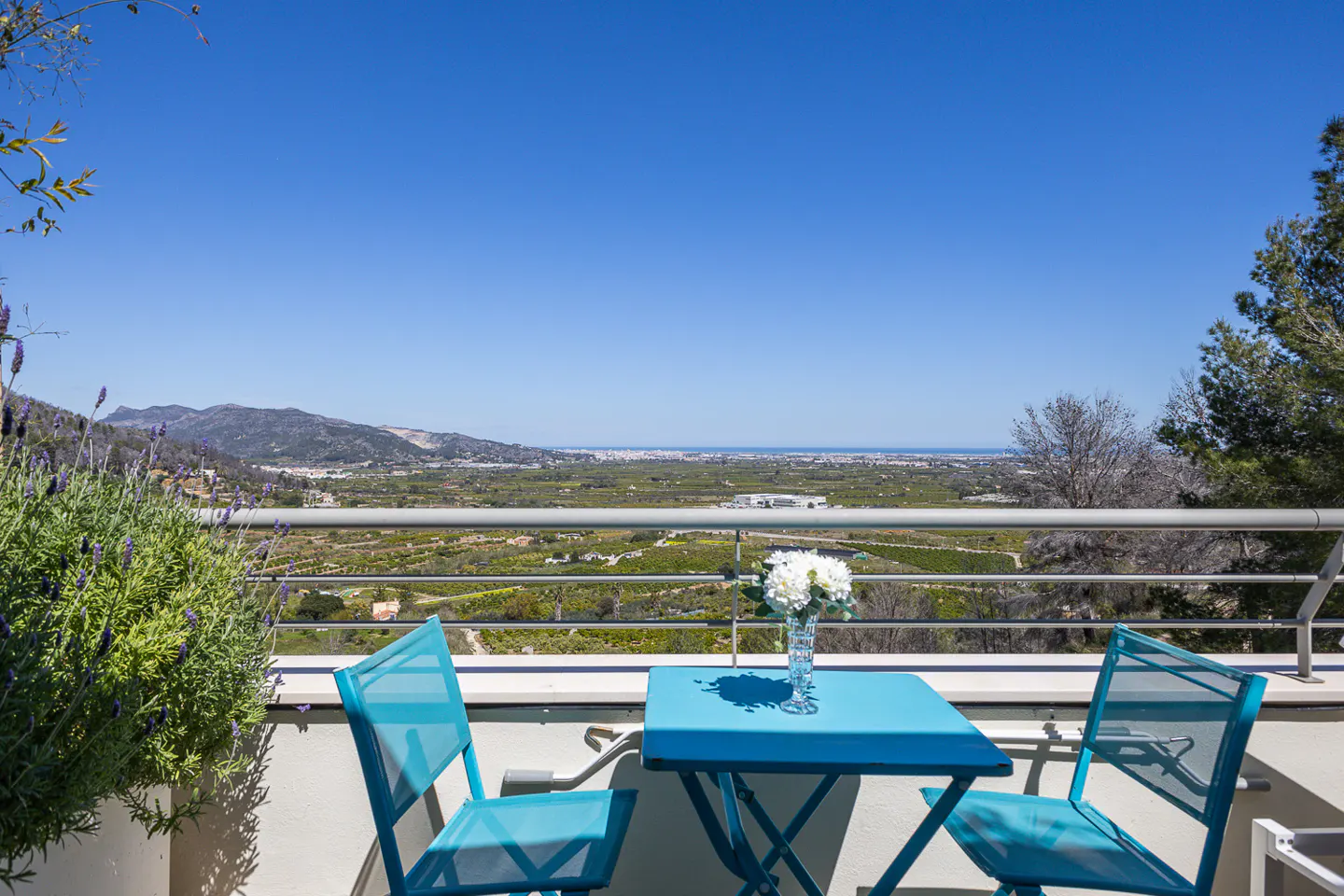 Balcony view with blue table and chairs, white flowers in vase, overlooking green landscape and mountains under a clear blue sky.
