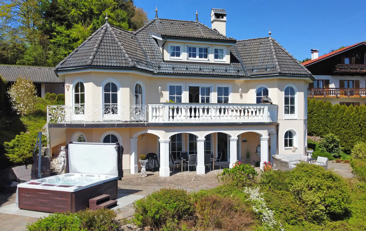 Exterior view of a two-story, light yellow house with a gray tiled roof, white balconies, a patio with outdoor furniture, and a hot tub.