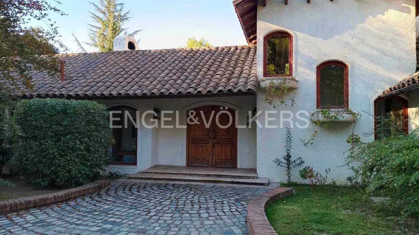 Exterior of a white stucco house with a brown tile roof and a cobblestone driveway. Engel & Völkers logo is visible.