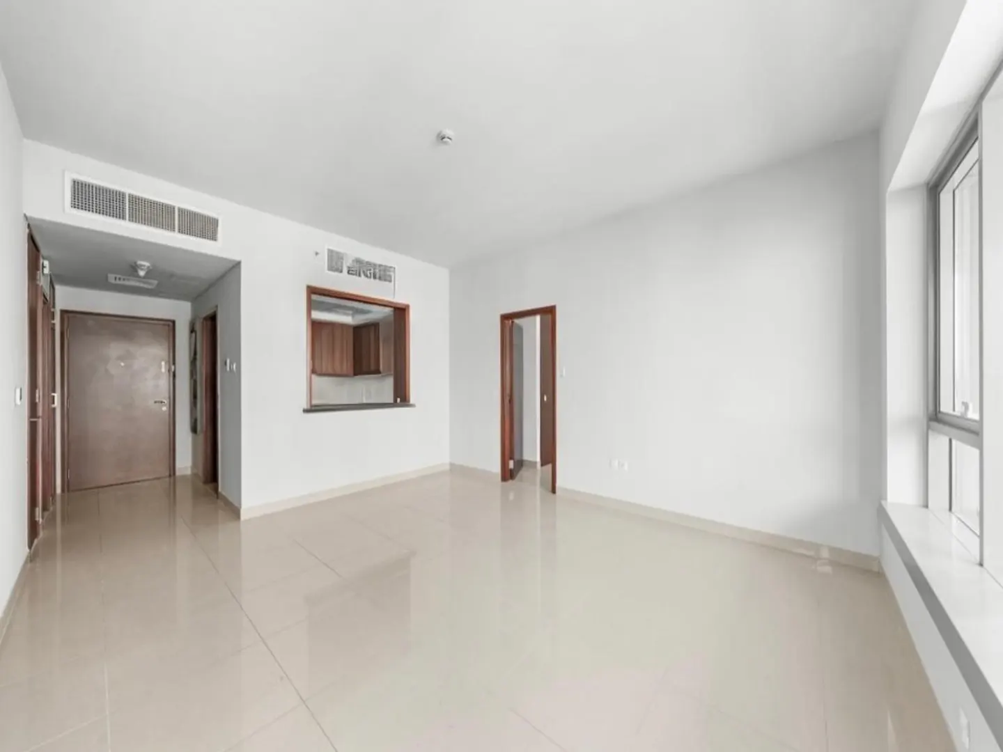 Bright, empty room with white walls and beige tile floor. A window lets in natural light. A doorway and kitchen pass-through are visible.