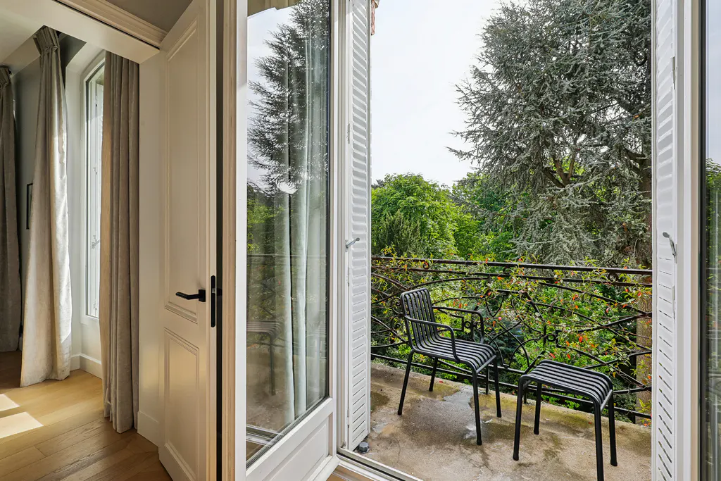 Open white French doors lead to a balcony with two black chairs and a view of lush green trees.