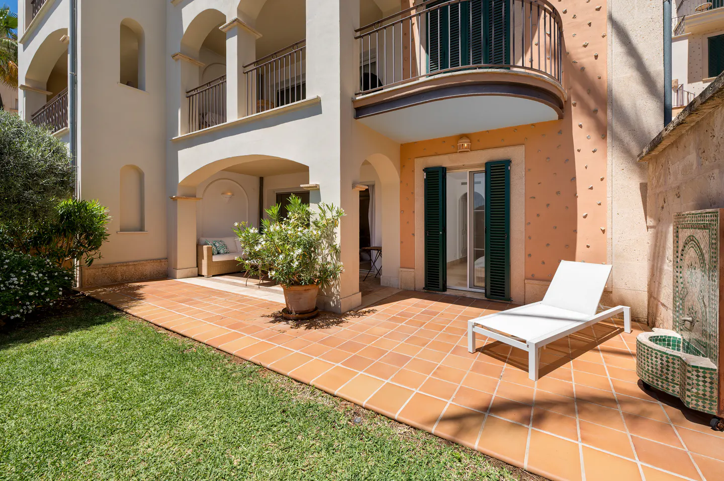 Exterior view of a building with a terracotta tiled patio, a white lounge chair, and a decorative fountain. Balconies with railings are visible above.
