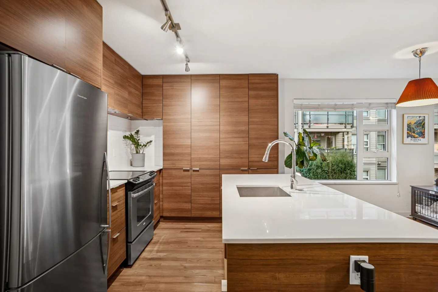 Modern kitchen with wood cabinets, stainless steel appliances, and a white countertop island. A window overlooks a green outdoor space.