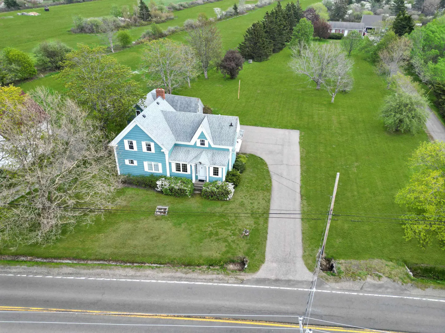 Aerial view of a light blue house with white trim, a gray roof, and a long driveway on a large green lawn.
