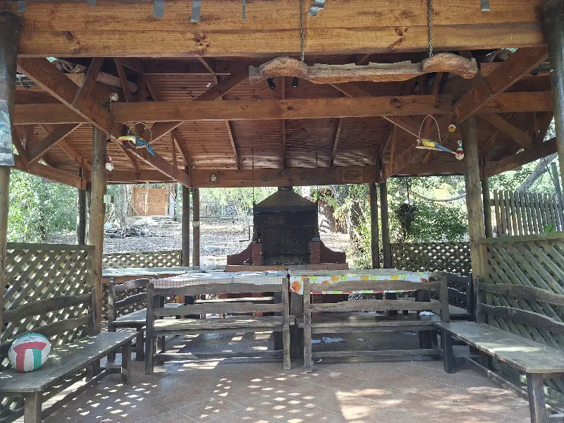 Outdoor wooden gazebo with benches, tables, and a grill. A volleyball sits on a bench. Trees are visible in the background.