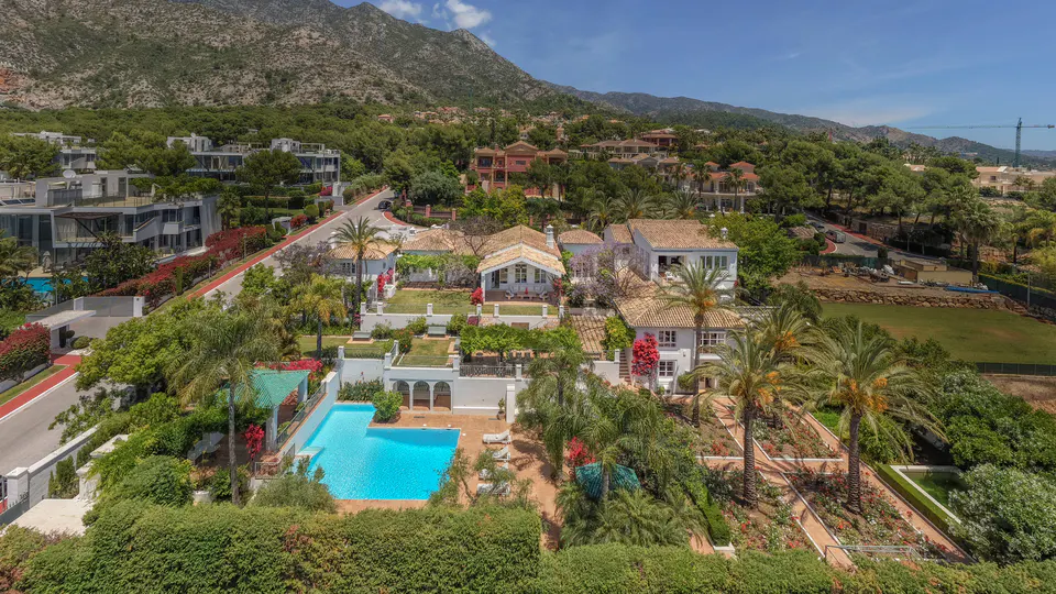Aerial view of luxury white villas with pools and palm trees, nestled in lush green hills under a clear blue sky.