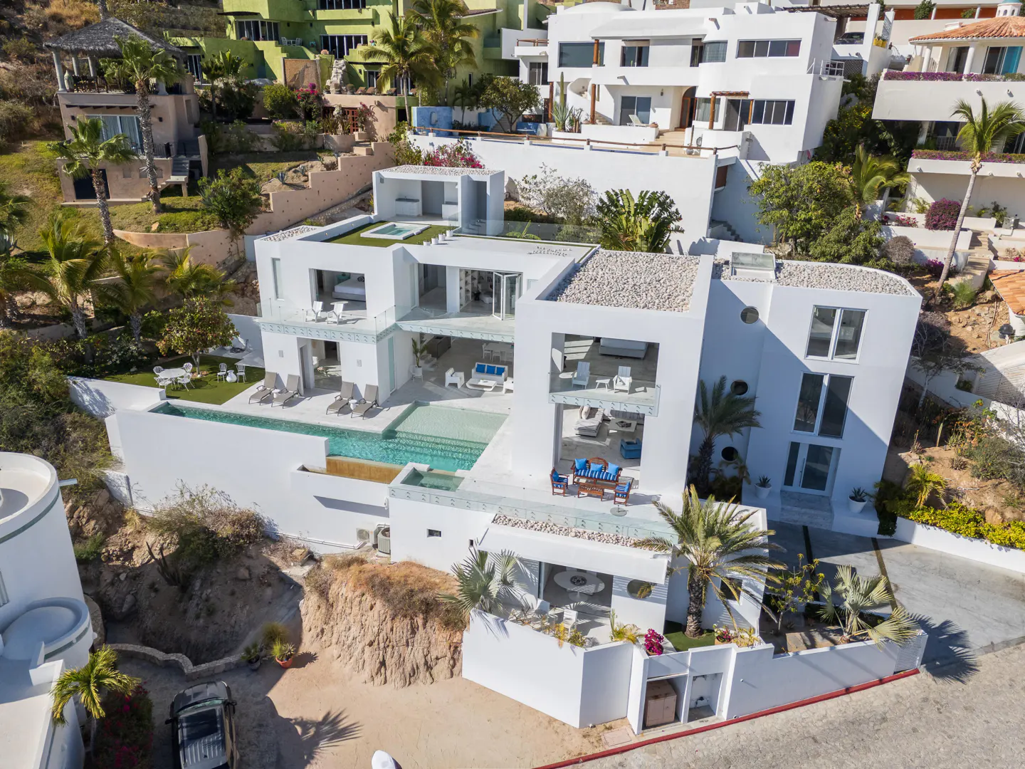Aerial view of a modern white multi-level villa with a pool, surrounded by lush greenery and other houses on a hillside.