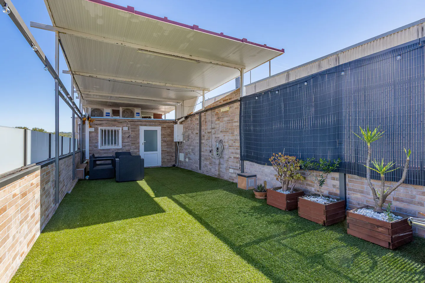 Outdoor patio with artificial grass, brick walls, and a covered roof. Potted plants line the back wall. A small seating area is visible.