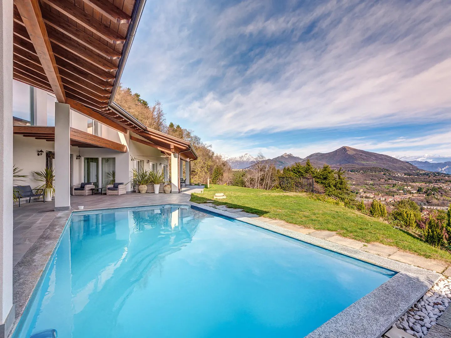A bright blue pool sits beside a modern white house with a wood-beam roof, overlooking a green hillside and distant mountains.