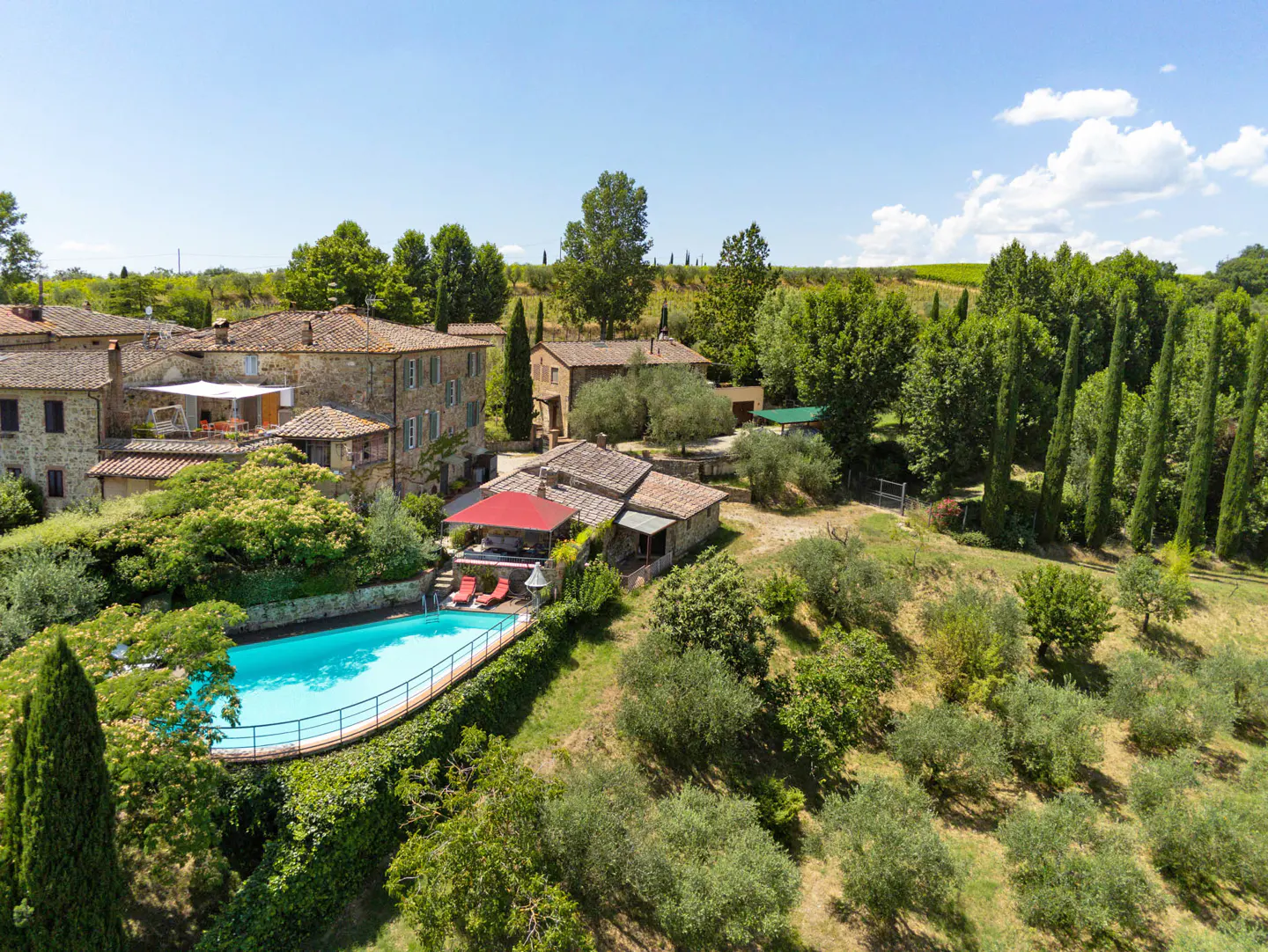 Aerial view of a Tuscan villa with a blue pool, red umbrellas, stone buildings, and green trees under a sunny sky.