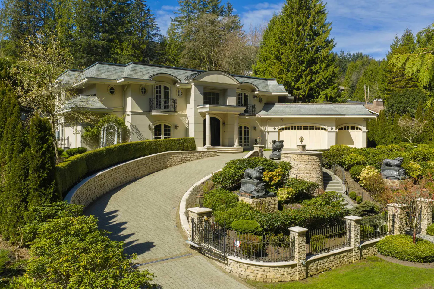 Beige two-story house with a gray roof, surrounded by green trees and bushes. A curved driveway leads to the house. Stone walls and black iron fences surround the property.