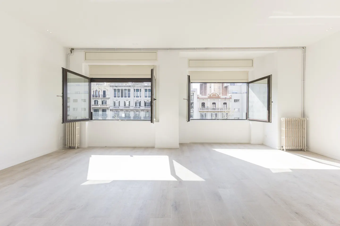 Bright, empty white room with two open windows showing city buildings. Radiators are under each window.