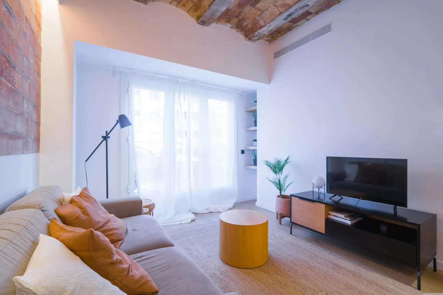 Living room with a beige sofa, orange pillows, a yellow table, and a black TV stand against a white wall.