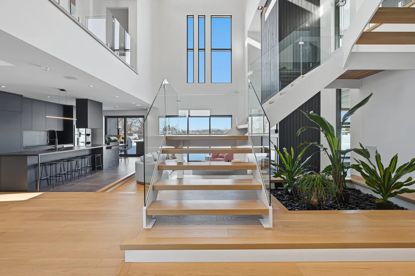 Bright, modern home interior with wood floors, a staircase with glass railings, and a black kitchen in the background.
