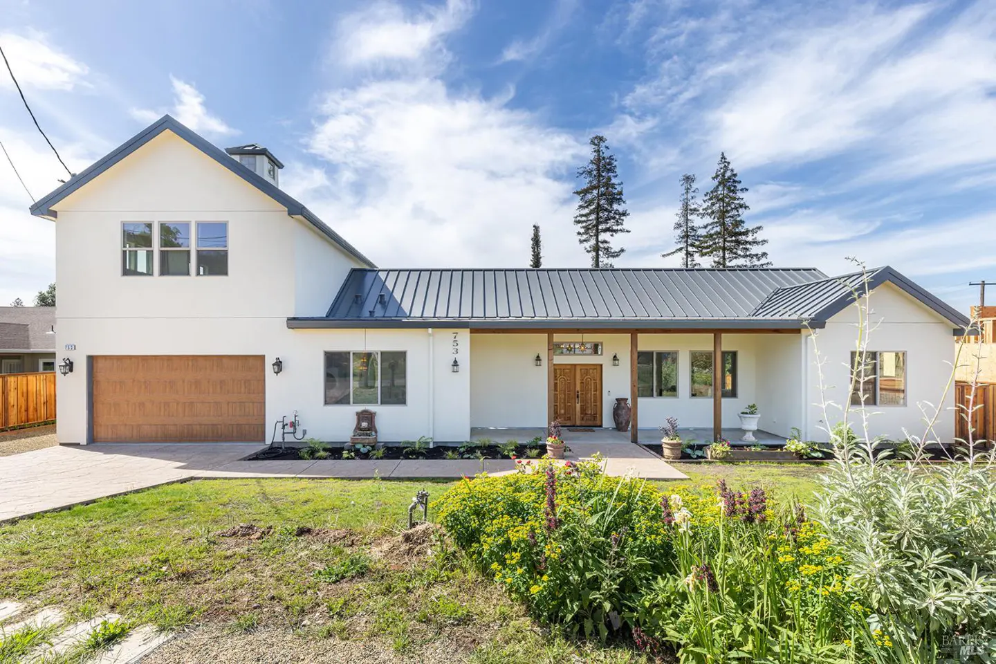 Two-story white house with a gray metal roof, brown garage door, and a front yard with flowers.