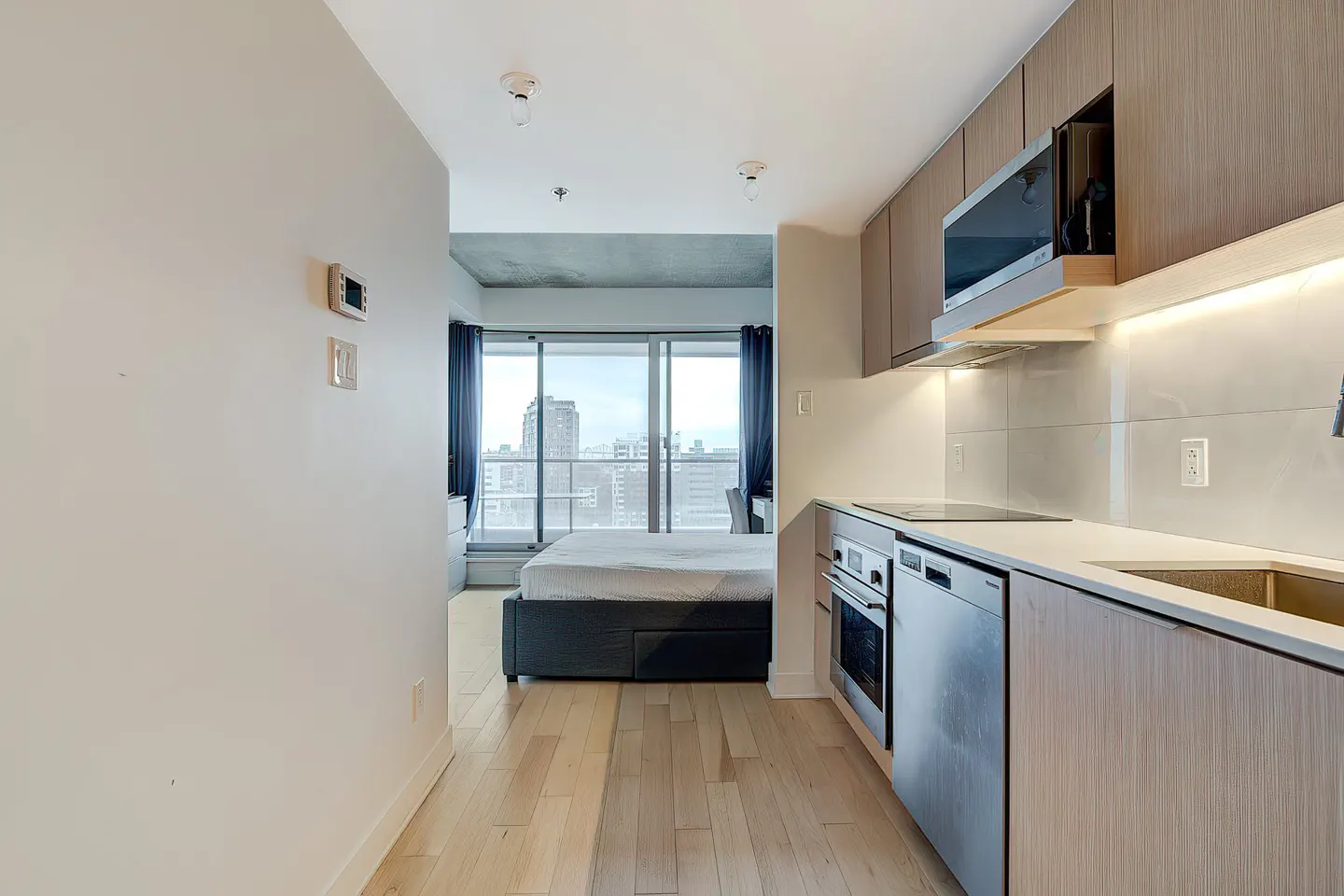 Studio apartment interior with a bed near a large window and a modern kitchen with light wood cabinets.