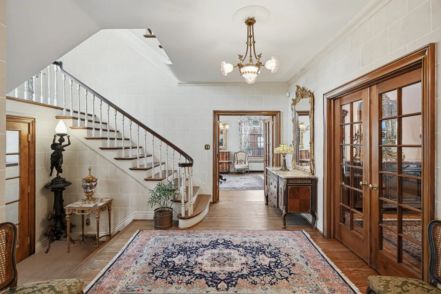 Elegant foyer with a staircase, wood floors, and a large patterned rug. A vintage chandelier hangs from the ceiling. French doors lead to another room.