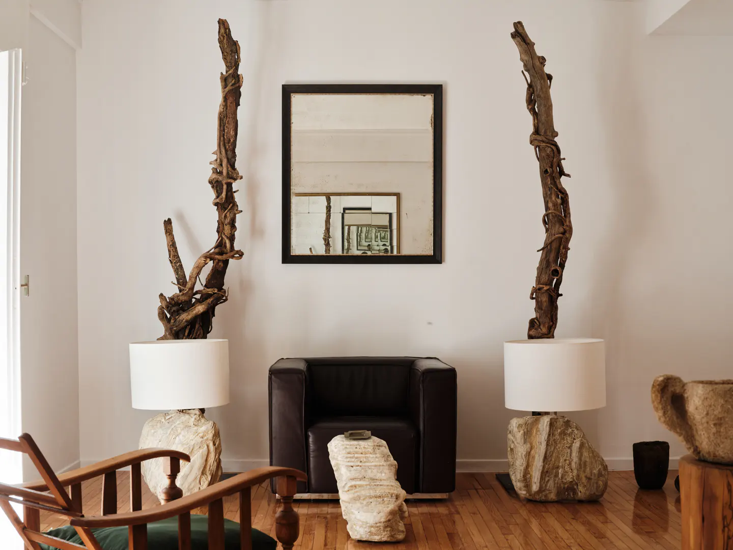 Living room with hardwood floors, white walls, a black leather chair, and a black framed mirror. Two lamps with driftwood and stone bases flank the chair.
