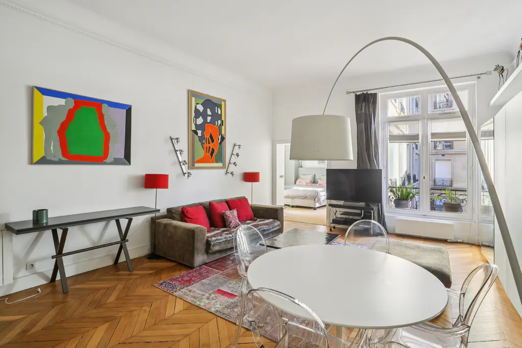Bright living room with parquet floors, white walls, and colorful art. A brown sofa with red pillows faces a white round table with clear chairs. A bedroom is visible in the background.